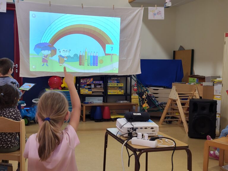 Momelino: Kinder im Klassenzimmer interagieren mit einer Momelino-Pr&auml;sentation &uuml;ber Farben und Formen auf einer Leinwand.