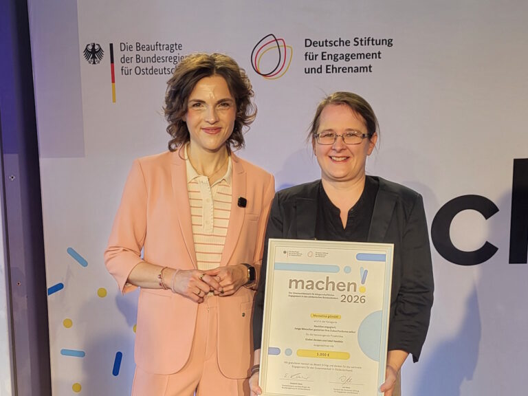 Momelino: Zwei Frauen stehen vor einer Wand mit Logos und halten eine Urkunde f&uuml;r den Ideenwettbewerb machen! 2026 f&uuml;r b&uuml;rgerschaftliches Engagement in die Kamera.